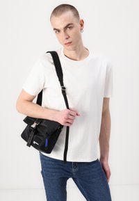 Crna torba za poruke s preklopom, s podesivim remenom, patentnim zatvaračem i odvažnim plavim naglascima. Nošena preko bijele majice i plavih traperica.