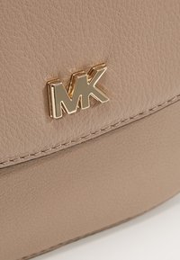 Vaaleanbeige nahkakäsilaukku, jossa on kultasävyinen "MK"-logo, teksturoitu pinta ja kontrastoiva ommel yksityiskohtana.