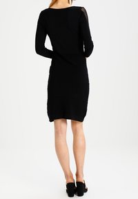 Robe noire à manches longues, corps texturé et accents transparents aux épaules. Présente une silhouette ajustée et tombe au-dessus du genou.