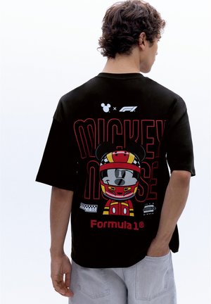 Homme portant un t-shirt noir avec Mickey Mouse en casque de course et des logos de Formule 1 dans le dos, tourné dos à la caméra.