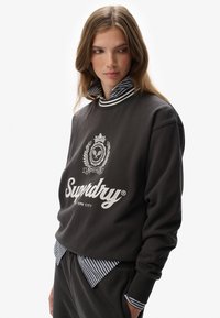 Charcoal sweatshirt with white logo and embroidered details, layered over a striped collared shirt. Soft fabric, relaxed fit, ribbed neckline.