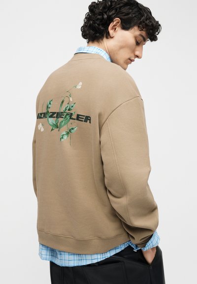 Νεαρός άνδρας με σγουρά μαλλιά, φοράει μπέζ φούτερ με πράσινο σχέδιο φύλλου και κείμενο στο πίσω μέρος, από πάνω από μπλε πουκάμισο με καρό μοτίβο.