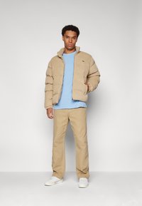 Beige puffer jacket with ribbed cuffs, light blue t-shirt, beige pants, and white sneakers. Features a logo on the jacket.