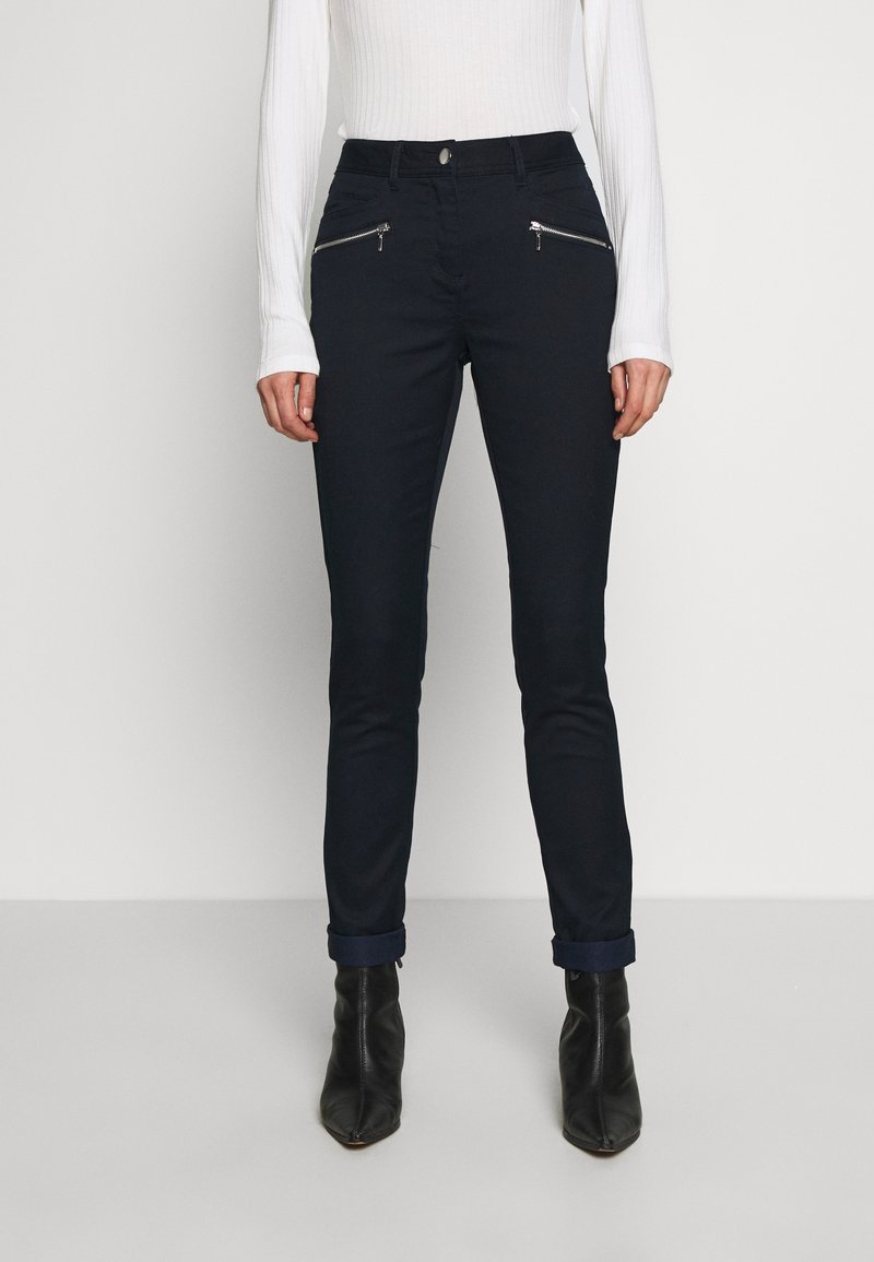 Femme portant un pantalon noir slim avec poches avant zippées, revers roulés, bottines noires et un top blanc à manches longues côtelé.