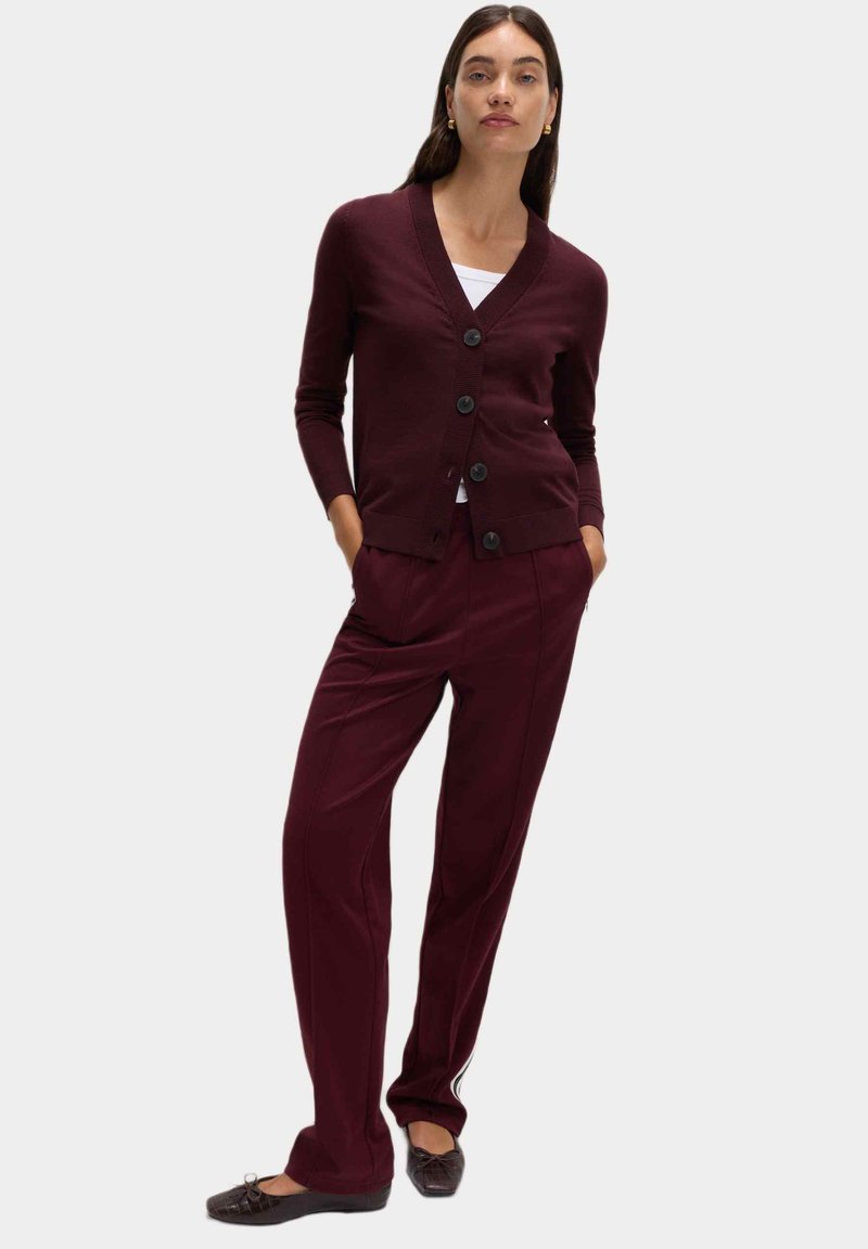 Kobieta ubrana w bordowy kardigan z guzikami i dopasowane spodnie, stojąca z rękami w kieszeniach, z jedną nogą lekko ugiętą.
