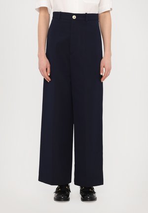 Pantalons larges bleu marine avec une texture lisse, dotés de plis sur le devant et d'une fermeture à bouton à la taille, associés à des chaussures noires brillantes.