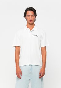 Polo shirt blanc avec une texture douce en coton, manches courtes et col. Présente un logo signature noir sur la poitrine gauche.