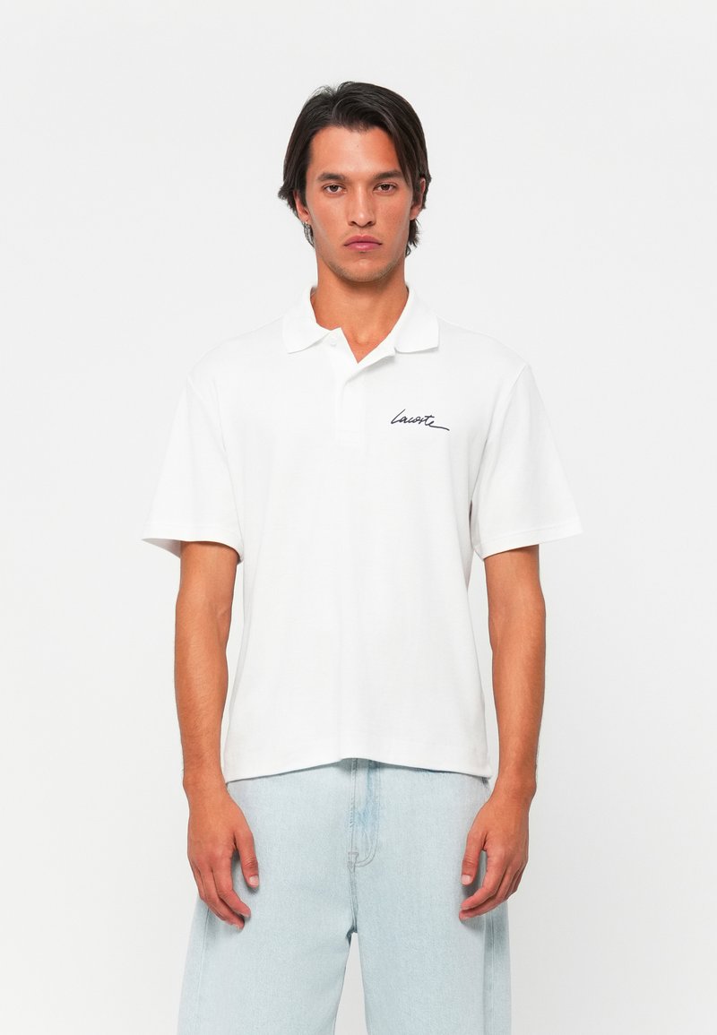 Polo shirt blanc avec une texture douce en coton, manches courtes et col. Présente un logo signature noir sur la poitrine gauche.