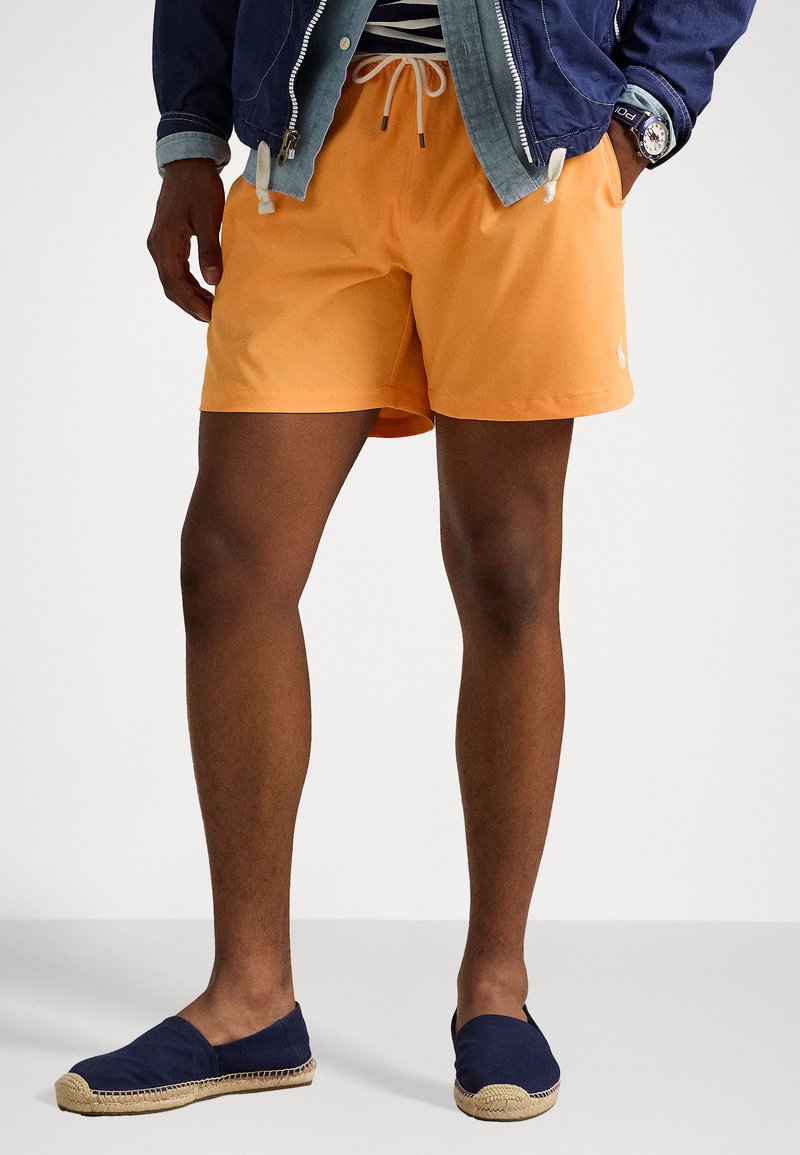 Bañador naranja con cintura ajustable por cordón, combinado con espadrilles azul marino que tienen una suela de yute trenzado. El conjunto se complementa con una chaqueta azul.