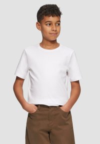 Jongen met krullend haar, gekleed in een eenvoudig wit T-shirt en bruine broek, staat met zijn handen in zijn zakken en kijkt opzij.