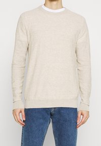 Beiger Rundhals-Pullover aus weichem Strickstoff, mit langen Ärmeln und gerippten Bündchen sowie Saum, getragen über einem weißen T-Shirt, kombiniert mit blauen Jeans.