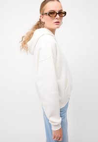 Hoodie bianco oversize con grande tasca frontale e cappuccio con cordino, abbinato a jeans azzurri chiari e occhiali da sole tartaruga.