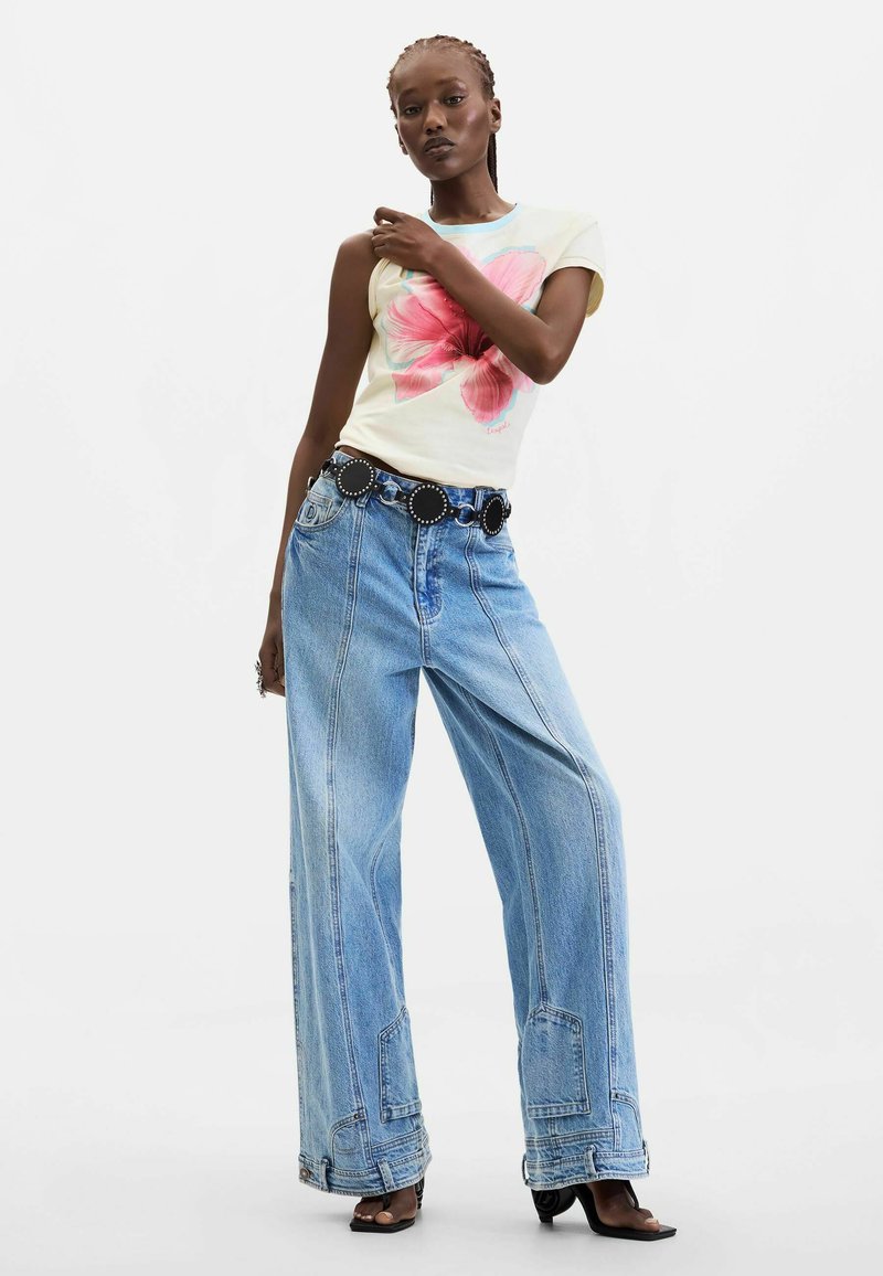 Woman standing in wide-leg blue jeans, black belt with large round buckles, and a white t-shirt featuring a large pink flower print.