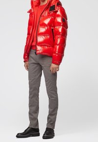 Chaqueta acolchada roja con superficie brillante, capucha y detalles en negro. Combinada con un suéter rojo, pantalones grises y zapatos negros. Ajuste casual.