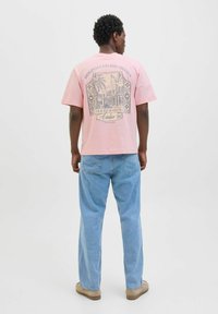 Rosa bomulds T-shirt med et trykt grafisk design på ryggen med arkitektonisk design, parret med lyseblå denimjeans og beige sko.