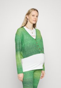 Cardigã em gradiente verde com decote em V, mangas longas e botões de madeira. Usado sobre uma blusa branca e combinado com leggings verdes iguais.