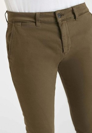 Close-up van olijfgroene broek met ceintuurlussen, zijzakken, knoopsluiting en een witte blouse die in de taille is gestoken.