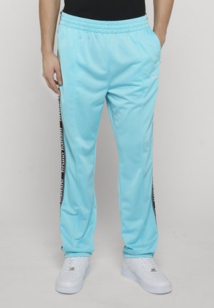 Person iført lyseblå treningsbukser med svarte og hvite stripete sidestriper med merke og hvite joggesko, stående mot en ensfarget bakgrunn.