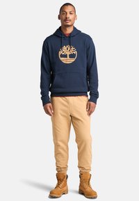 Granatowy hoodie z złotym logo drzewa, kieszenią kangurką, w zestawie z tanowymi spodniami dresowymi i brązowymi wiązanymi butami.
