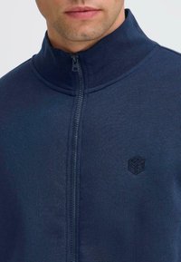 Tamsiai mėlyna zip-up striukė su aukšta apykakle, lygia tekstūra ir mažu juodu logotipu kairėje pusėje. Užtrauktukas eina vertikaliai per priekį.