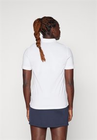 Weißes Poloshirt mit kurzen Ärmeln, gestaltet mit einem Kragen und einer schmalen Passform. Dazu gehört ein marineblauer Rock und geflochtenes Haar, das nach hinten gebunden ist.