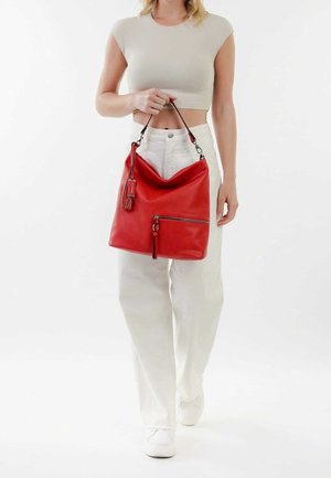 Femme portant un pantalon blanc et un crop top, tenant un grand sac à main en cuir rouge avec une poche à fermeture éclair et des détails en pampilles.