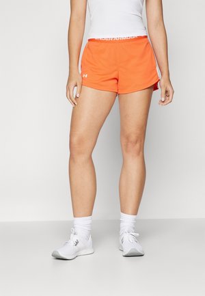 Persona che indossa pantaloncini sportivi arancioni Under Armour, canottiera bianca, calzini bianchi e scarpe sportive bianche, in piedi davanti a uno sfondo semplice.