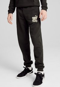 Pantalones de chándal negros hechos de una tela suave, que presentan un gráfico blanco "PUMA PARK SQUAD" en el muslo izquierdo y puños elásticos en los tobillos.