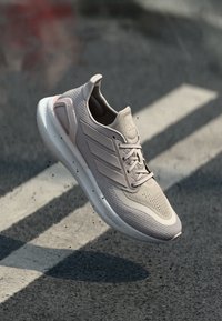 Chaussure de sport gris clair avec une tige en mesh, un design en tricot texturé, des accents à trois bandes et une semelle rembourrée. Flottant au-dessus d'une surface texturée.