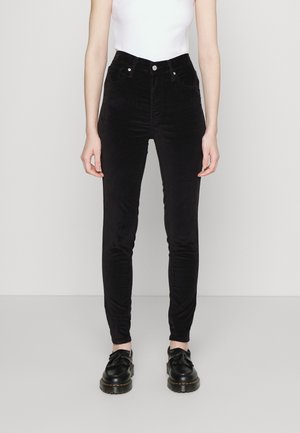 Pantalon slim en velours noir porté avec des mocassins noirs à pampilles et un top blanc sans manches, modèle debout devant un fond uni.