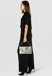 Metallisch silberne Clutch mit Kettenhenkel, kombiniert mit einem schwarzen, plissierten Overall und spitzen schwarzen Pumps. Seitenansicht.