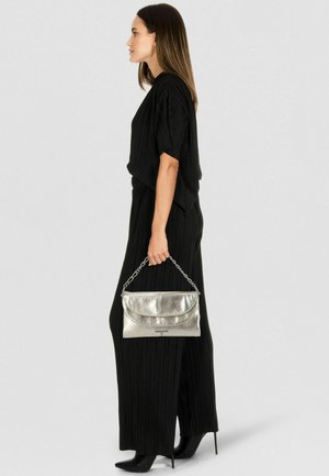 Bolso de mano plateado metálico con asa de cadena, combinado con un mono negro plisado y tacones negros puntiagudos. Vista de perfil lateral.