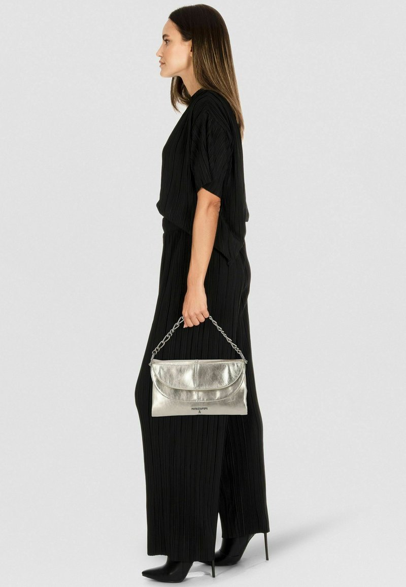 Metallisch silberne Clutch mit Kettenhenkel, kombiniert mit einem schwarzen, plissierten Overall und spitzen schwarzen Pumps. Seitenansicht.