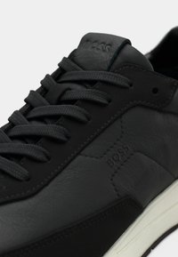 Μαύρο δερμάτινο αθλητικό παπούτσι με υφασμένες λεπτομέρειες, επίπεδα κορδόνια, λευκή σόλα και ανάγλυφο λογότυπο. Αντίθεση ανάμεσα σε λεία και ματ επιφάνεια.