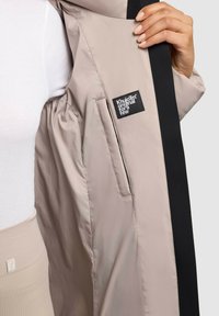 Chaqueta beige claro con un forro negro, con un parche de logotipo, textura suave y un bolsillo lateral bajo la solapa.