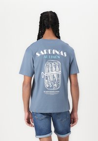 Blå bomulls t-skjorte med et retro sardinkonservesgrafikk på baksiden. Teksten sier "SARDINAS AL LIMÓN." Designet inkluderer fet skrift og detaljerte illustrasjoner.