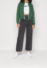 Vihreä vetoketjullinen takki valkoisella kantilla, yhdistetty valkoiseen crop-toppiin, jossa on vihreä raita. Mustat leveälahkeiset farkut ja valkoiset tennarit.