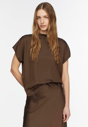 Młoda kobieta z długimi falującymi włosami, ubrana w luźny brązowy top z krótkim rękawem i pasującą spódnicę, stoi na jednolitym białym tle.