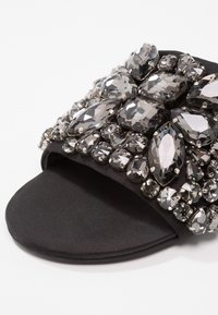 Sandale noire de type slide avec de gros cristaux clairs facettés embellissant la large bride supérieure sur fond blanc.