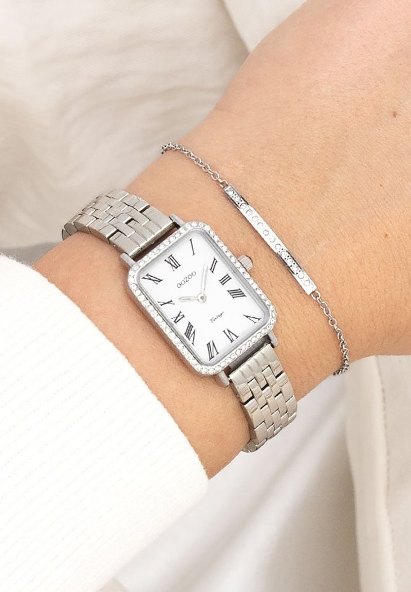 Montre-bracelet en acier inoxydable argenté avec un cadran rectangulaire, un fond blanc, des chiffres romains noirs et une lunette ornée de cristaux sur un bracelet tressé.