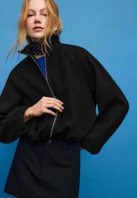 Sort lynlåsjakke med høj krave og voluminøse ærmer, lavet af tekstureret stof. Kombineret med en marineblå nederdel, mod en blå baggrund.