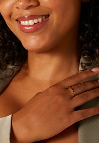 Gouden ring met een gestructureerde band gedragen aan een hand; een gladde huidtint en een kledingstuk met kraag zijn zichtbaar in het beeld. Een stralende glimlach versterkt de scène.