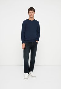 Pull en maille côtelée bleu marine avec un col ras du cou, associé à un jean foncé et des baskets blanches. Le modèle se tient les mains sur les côtés devant un fond blanc.