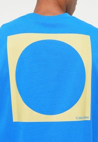 Mėlynas medvilninis marškinėliai su dideliu geltonu kvadratu ir apvaliu išpjovimu nugaros pusėje, apačioje esantis "Calvin Klein" logotipas.