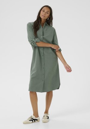 Groen linnen shirtjurk met lange mouwen, knoopsluiting aan de voorkant en zijsplitten. Gecombineerd met beige sneakers met zwarte accenten.
