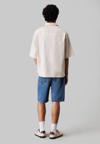 Korte mouwen, lichtbeige shirt van zachte stof, gecombineerd met blauwe denimshorts. Sneakers met witte sokken maken de outfit compleet.