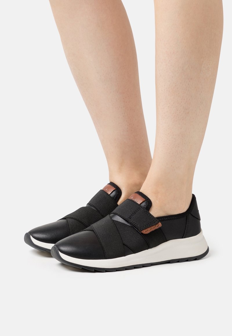 Pieds portant des baskets noires sans lacets avec des brides croisées élastiques, des étiquettes de logo marron et des semelles blanches sur un fond clair uni.