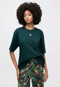 Mørkegrøn bomulds t-shirt med korte ærmer, med et lille hvidt Adidas-logo på fronten. Kombineret med flerfarvede bukser med blomsterprint.