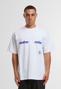 Hvid bomulds T-shirt med korte ærmer. Har blå tekst, der står "another" og "cotton" på forsiden, samt et blåt logo.