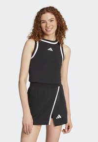 Crop top de sport noir avec des accents et un logo blancs, associé à une jupe noire avec une rayure blanche et une fente latérale. Texture de tissu douce.